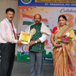 Man in a pink shirt presents a certificate to a man in a green kurta while a woman in a sari holds a bouquet on stage